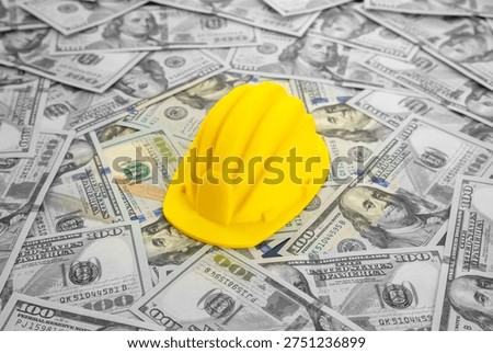 A yellow construction worker's hard hat against a background of money. Symbol of booming construction sector, real estate bubble risks and major investments in building projects, engineering