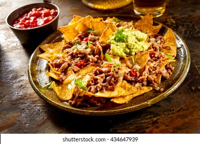 Yellow corn nacho chips garnished with ground beef, guacamole, melted cheese, peppers and cilantro leaves in plate on wooden table - Powered by Shutterstock