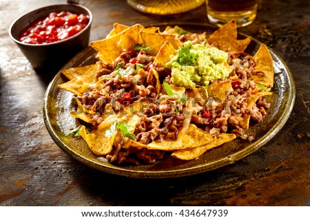 Yellow corn nacho chips garnished with ground beef, guacamole, melted cheese, peppers and cilantro leaves in plate on wooden table