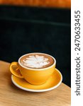 A yellow cup of hot sweet mocha coffee with latte art on top on a saucer on the edge of a wooden table in a cafe, copy space