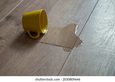 Yellow Cup Of Water Spilled On Wooden Laminate Floor. Moisture Protection Of A Floor. Parquet Board With Spilled Water.