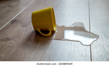 Yellow Cup Of Water Spilled On A Wooden Laminate Floor. Moisture Protection Of A Floor. Waterproof Parquet Board With Spilled Water.