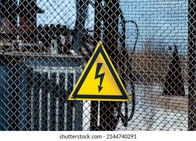 Yellow electricity warning sign with lightning hanging on gray wire mesh fence that surrounds transformer.