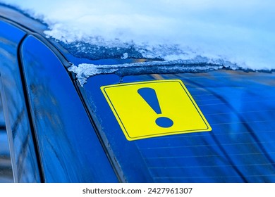 Yellow exclamation mark in selective focus on the rear window of a car with snow in winter. Designation of an inexperienced driver behind the wheel, a warning signal.
