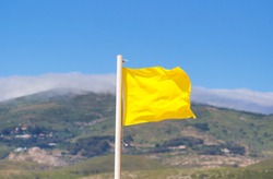 Yellow Flag Waving On The Beach In The Breeze Against A Blurred Blue Sky.