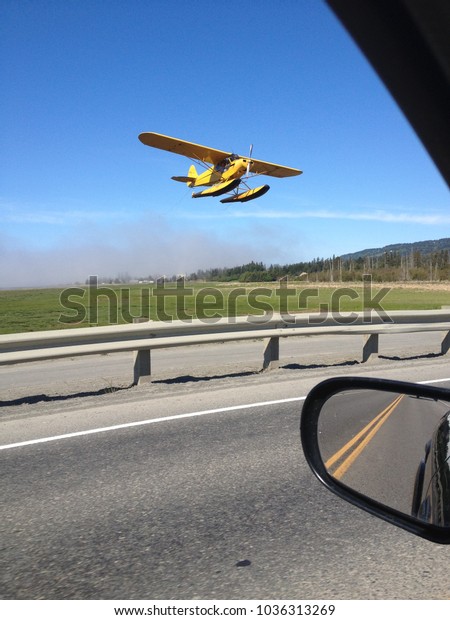 Yellow Float Plane Flying over road\
and car coming into land on Beluga Lake in Homer\
Alaska