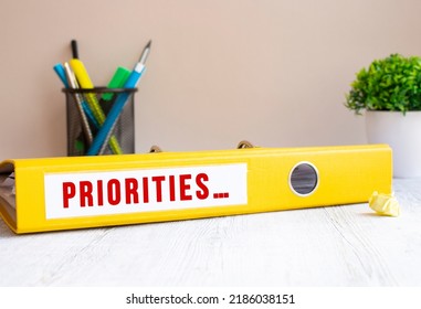 A Yellow Folder Labeled PRIORITIES Is On The Office Desk. Flower And Stationery Background. Financial Concept.