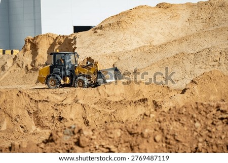 Yellow front loader moving sand near large soil embankment and rough excavation cut for construction logistics and site preparation industry