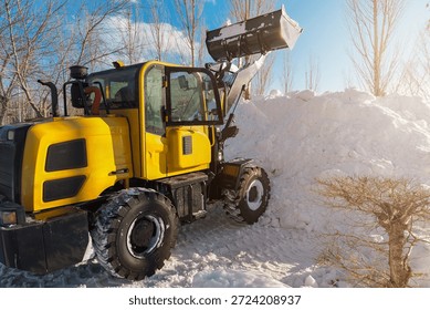 Yellow front-end loader clearing a large pile of fresh snow on a sunny winter day. Heavy equipment performing essential snow removal, showcasing powerful machinery against a clear blue sky outdoors.