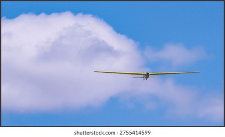 Yellow glider plane soaring through a bright blue sky with soft white clouds, captured from a distance during a graceful recreational flight on a sunny day.