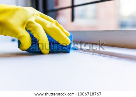 Yellow gloves hand cleaning dust, dirt on windowsill with blue sponge by window in urban apartment in New York City NYC