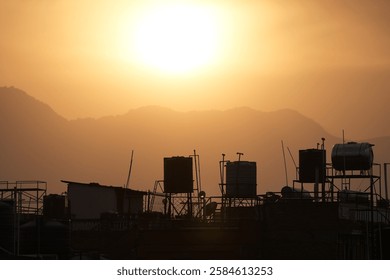 El resplandor amarillo y la niebla de la hora dorada del atardecer se proyectan sobre la densa capital urbana de Katmandú, Nepal. Los tanques de agua domésticos en los techos forman una silueta geométrica contra un fondo montañoso.
