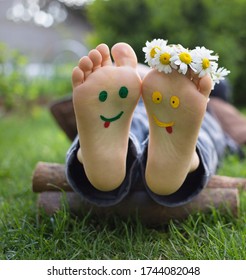 Yellow And Green Faces Are Painted On The Bare Feet Of A Child Lying On The Grass. Chamomile Flowers, Green Grass, Joy, Cheerful Positive Atmosphere, Happy Childhood. Hello Summer, Smile Day