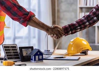 Yellow hard hat on table and house model solar windmill with construction team handshake to start new project contract plan in renewable energy concept contractor office center - Powered by Shutterstock