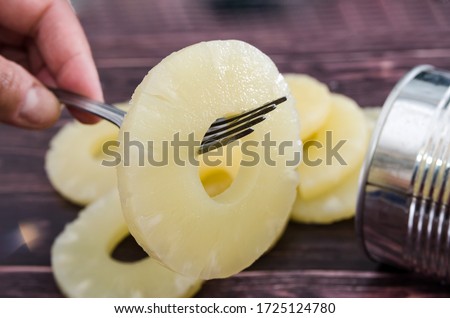 Yellow juicy pineapple ring on a fork against the background of a tin can. Caramelized canned pineapple. Raw food diet. Close-up plan. Woody background.