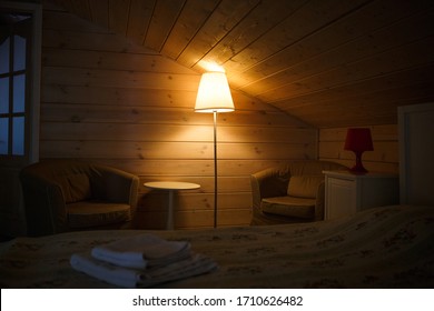 A Yellow Lampshade Shines Dimly And Comfortably In The Attic Bedroom With A Low Ceiling. Lamp Near The Bed. Interior Of A Wooden Bedroom In A Country House.