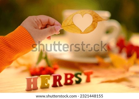 yellow leaf with heart in female hand, tea, coffee in a mug on the table in the garden, colored letter words, the concept of outdoor tea drinking, good weather, a cozy autumn mood