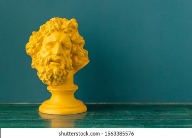Yellow male head with beard and long hair of the ancient Greek sculpture on a floor against blue wall background. Hellenistic Period 323 BCE – 31 CE