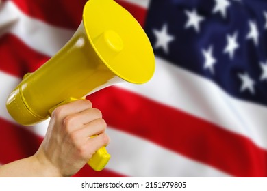 A yellow megaphone in a man's hand against the background of the national flag of the United States of America. False rumors, fakes about the state, country, elections, debates of candidates. - Shutterstock ID 2151979805