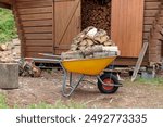 Yellow metal garden wheelbarrow and a pile of sawn firewood near the firewood storage. Preparing firewood for the winter to heat the house