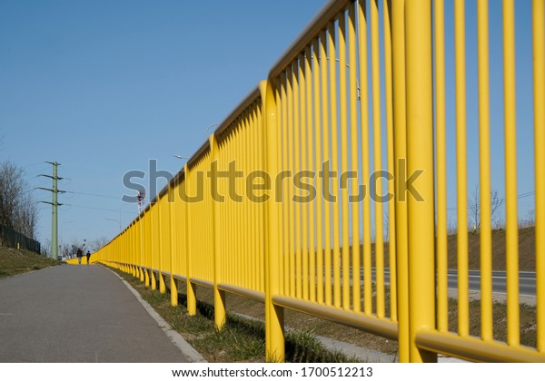 Yellow metal safety barriers along\
the street. Silhouettes of walking people on\
pavement.