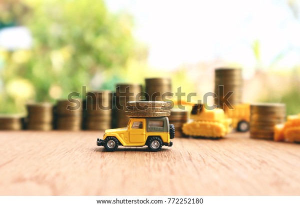 Yellow\
miniature car carry Dollar Singapore coins and blur construction\
vehicles help put coins into rolls ladder of gold money on wood\
table in blur natural tree bright\
sunlight