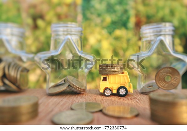 Yellow miniature truck carry Dollar Singapore coins\
and star shape bottle with blur gold money on wood table in blur\
natural tree