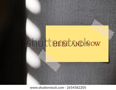 Yellow note on wall with text written HERE AND NOW - refers to present moment, being conscious and mindful of what happening right now than on past or worrying about  future