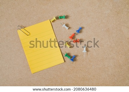 yellow note paper with paper clip and multi color thumbtacks on brown grunge board, top view