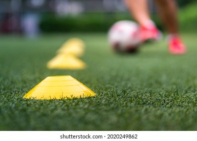 A yellow obstacle cone (Focus), sport training equipment on artificial pitch ground with action of a football player is practice to dribbling the ball as blurred background. Sport training concept. - Powered by Shutterstock