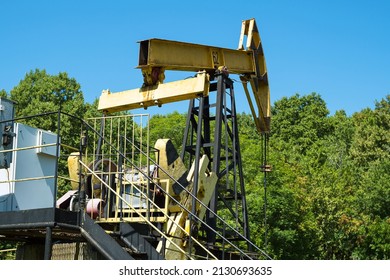 A Yellow Oil Rig Pumping Oil From A Well Against A Backdrop Of Green Trees And Blue Sky. Side View From Behind