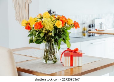 Yellow And Orange Roses In A Vase And Gift Box With Red Ribbon On Kitchen Counter Table With Modern Kitchen Background. Gift For Thanksgiving Day, Birthday, Mother's Or Valentine's Day. Copy Space