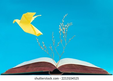 Yellow origami dove flying from an old book on blue background. Soft focus close up of a dry tree growing from a book