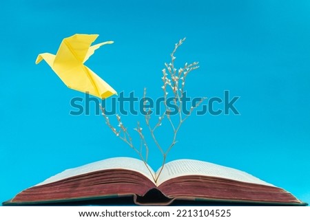 Yellow origami dove flying from an old book on blue background. Soft focus close up of a dry tree growing from a book