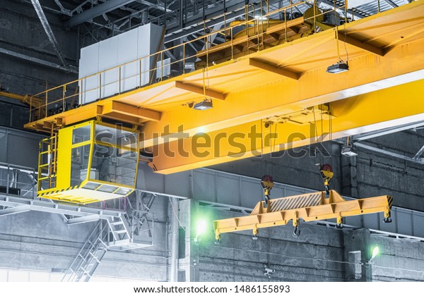 Yellow overhead crane with linear traverse and\
hooks in engineering plant shop. Cabin of crane operator and jib\
crab trolley. Landing \
staircase.