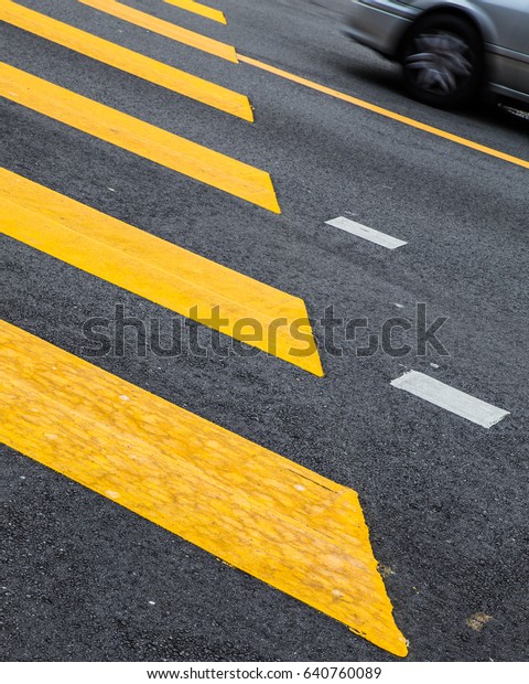 Yellow painted crosswalk road line markings\
with moving car in\
background