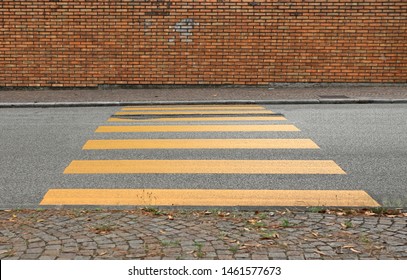Yellow Painted Crosswalk Road Lines On Asphalt Between Two Porphyry Sidewalk. Grunge Brick Wall On Background. Suburban Background For Copy Space