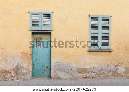 Yellow painted house wall with cracked surface, two windows and green metal door, street view