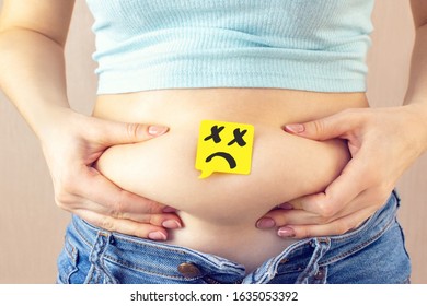A yellow paper blank message is drawn: a sad face on belly fat with hands squeezes at the waistline of a young woman in jeans. The concept of excess weight, weight loss, diet, obesity, junk food. - Shutterstock ID 1635053392