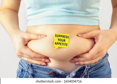 A yellow paper blank message is written Suppress your appetite on belly with hands squeezes at the waistline of a young woman in jeans. The concept of loss excess weight, diet, obesity, junk food. - Shutterstock ID 1654034716