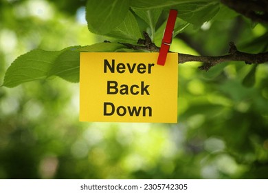 A yellow paper note with the phrase Never Back Down on it attached to a tree branch with a clothes pin. Close up.