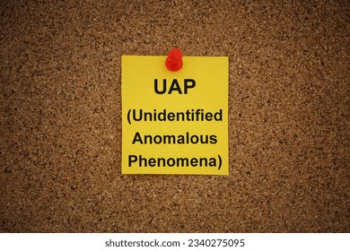A yellow paper note with the words UAP (Unidentified Anomalous Phenomena) on it pinned to a cork board. Close up.