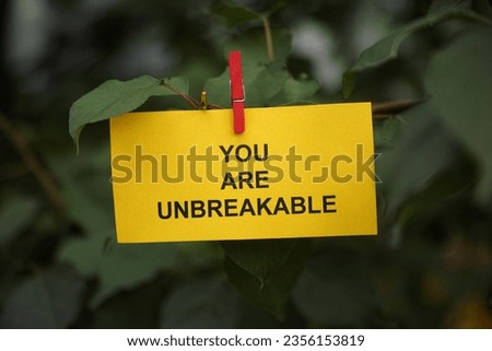 A yellow paper note with the words You Are Unbreakable on it attached to a tree branch with a clothes pin. Close up.