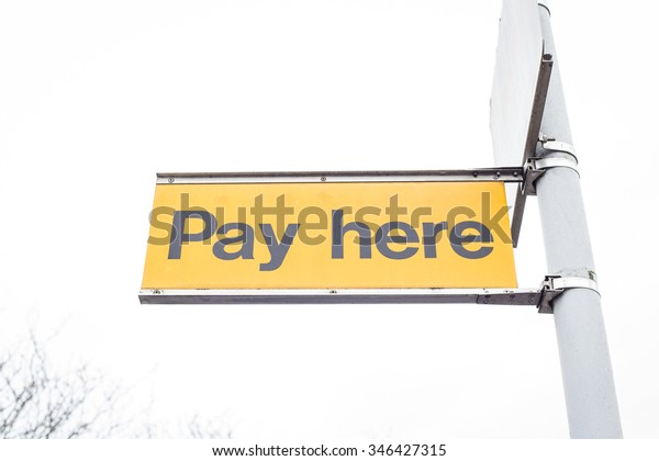 Yellow pay here car parking sign on a white\
sky background.