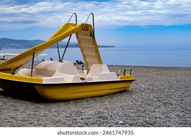 Yellow pedal boat with slide on a pebbly beach, serene summer vacation spot – calm sea and partly cloudy sky – copy space. - Powered by Shutterstock