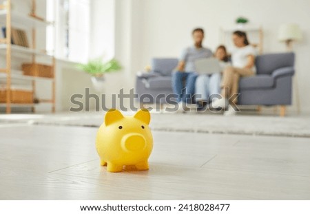 Yellow piggy money box standing on floor in the living room and a parents with a child girl sitting on sofa at home in background. Family savings, investment and money management concept.