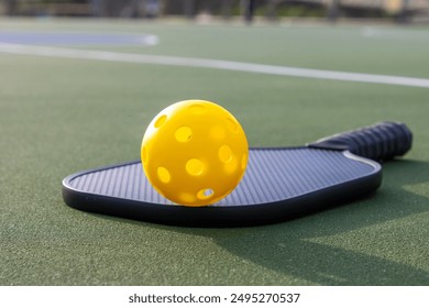 Yellow plastic pickleball ball and a black paddle on an outdoor playing court.