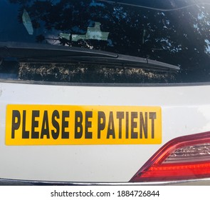 A Yellow Please Be Patient Sticker On A White Car Of A Student Driver. The Rear Windshield And Taillight Can Also Be Seen.