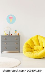 Yellow Pouf Next To A Grey Chest Of Drawers And Ice-cream Poster On A White Wall In Kid Room Interior. Place For Your Poster