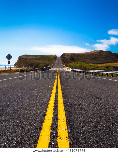 Yellow road dividing lines from low\
perspective along coastal road leading towards\
hills